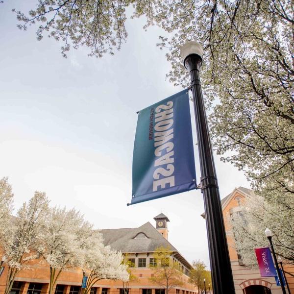 A Reach Higher Showcase banner hangs from a post outside the DeVos Center, surrounded by blossoming trees.