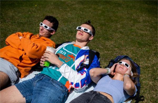 Three people lie on the ground while wearing special glasses for an eclipse. They are looking skyward.