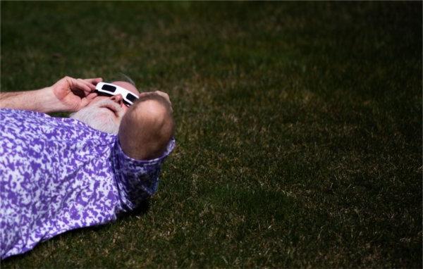 A person lying on the ground holds special glasses against their head while viewing the solar eclipse.