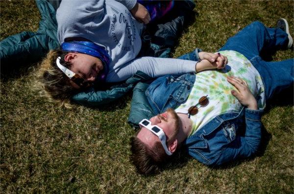 Two people hold hands while lying on the ground and wearing special glasses to view the solar eclipse.